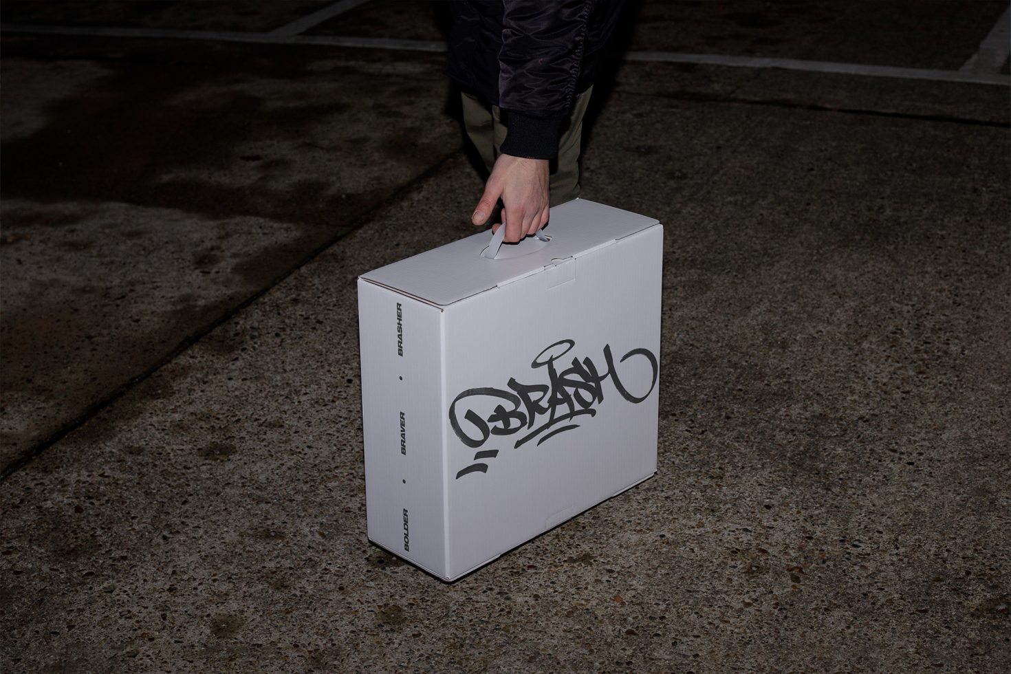 Person holding a white box with black graffiti-style logo, urban setting, for use in mockups, packaging design, branding assets.